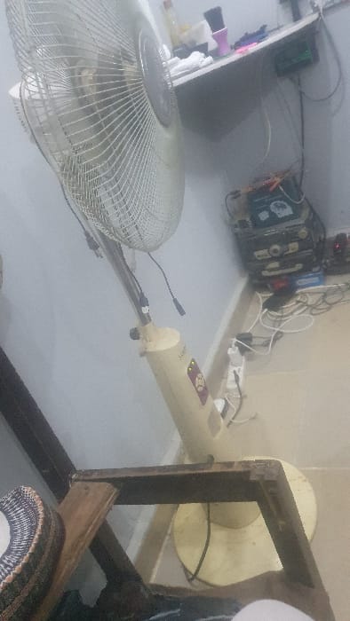 The image displays an indoor scene located in the city of Danbare, Nigeria. A cream-colored pedestal fan with a metallic grille and a visible "AHSAN" logo on its base stands on a light-colored tiled floor, positioned in the center-right of the frame. Its black power cord connects to a white power strip, which is situated on the floor amidst a tangle of other electrical cables. To the right of the fan, a stack of dark-colored electronic equipment, including one unit with a visible circuit-like diagram, rests on the floor.

Above the electronic equipment, a white shelf is mounted on a light blue wall. Items on the shelf include a clear bottle, folded white fabric, a purple and black brush, and a dark blue object. A small green digital display is visible on the right side of the shelf, connected by wires. In the bottom-left foreground, a dark wooden frame, possibly part of a table or chair, is partially visible. Next to the wooden frame, a stack of patterned fabric items, resembling multiple hats, is present. No people are visible, and no active interactions or actions are depicted.