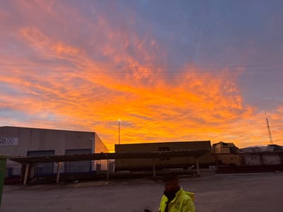 The image captures an industrial area within Polígono Industrial Nicomedes García, Spain, at either sunrise or sunset, characterized by a sky displaying intense orange, red, and yellow hues with wispy, stratified clouds, transitioning to a lighter orange and purple-grey near the top. Multiple thin, dark horizontal lines, consistent with power lines, traverse the upper sky.
In the lower right foreground, a male subject with dark hair and beard is partially visible, facing left. He is wearing a high-visibility yellow-green jacket over a dark undergarment and appears to be holding a small electronic device.
The ground surface is a grey, paved area. A tall street lamp with an illuminated yellow-orange light source is positioned in the middle-right.
To the left, a large, light-colored industrial building with "SDLOG" visible on its side features multiple loading docks with dark roller doors beneath a dark horizontal canopy structure. A green dumpster is partially visible on the extreme left.
A dark, elevated canopy structure spans horizontally across the midground. Behind this, a long yellow shipping container or trailer, marked with a logo resembling "M" or "MO," is parked. Further right, several articulated lorries, at least one with a yellow cab, are present. In the far right background, industrial structures and a tall, dark communication tower with an antenna are visible.