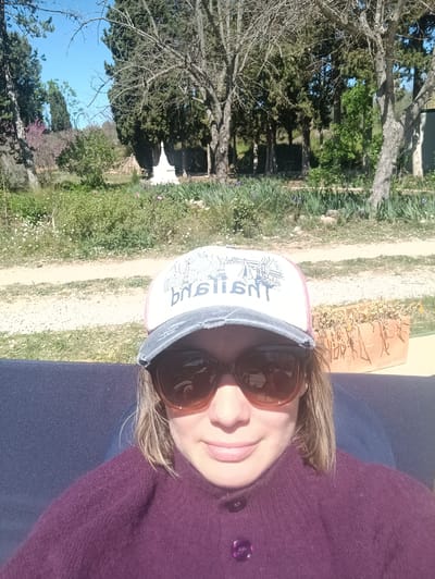 A woman wearing sunglasses and a cap with the word "basilica" (possibly Basilica de la Virgen de los Desamparados) sits outdoors. She is wearing a purple sweater. The background includes trees, grass, and a path. A small white structure is visible further back. The location is likely Jérica, Spain.
