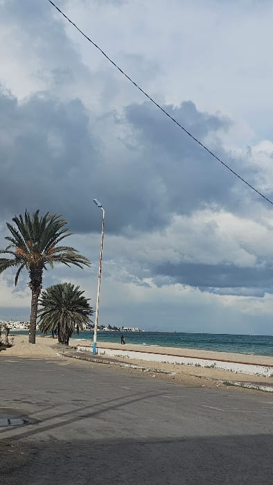 The image depicts a coastal scene in Sousse, Tunisia, under an overcast sky with heavy grey clouds dominating the upper two-thirds of the frame. A dark utility cable runs diagonally from the upper left to the upper right corner. In the foreground, an asphalt road with visible cracks and minor depressions occupies the left side, featuring long, dark shadows extending from left to right across its surface.

Adjacent to the road, a low, light-colored concrete barrier separates it from a sandy beach. Several palm trees with dense green fronds are positioned on the sand to the left and center-left, behind the barrier. A tall, slender street lamp with a light blue fixture and a weathered, discolored pole stands between the road and the beach, slightly right of the prominent palm trees.

Beyond the beach, the Mediterranean Sea exhibits turquoise water with visible wave activity. A single individual, dressed in dark clothing, walks along the water's edge on the beach, facing away from the viewer and moving towards the right. In the far distance, across the sea, a low-lying urban area with numerous white buildings is visible, suggesting a distant part of Sousse or its environs. A faint structure, possibly a lighthouse or pier, extends into the sea on the distant horizon to the right. The overall lighting suggests daylight under cloudy conditions.