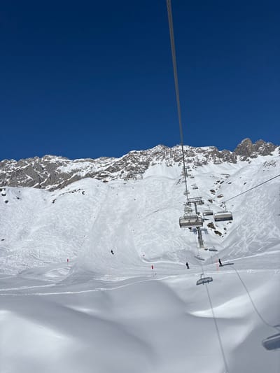 A high-angle photograph captures a sunlit, snow-covered mountain landscape at a ski resort in Klösterle, Austria. The foreground and midground feature extensive ski slopes with numerous ski tracks on pristine white snow, interspersed with undulations creating shadowed areas. A prominent ski lift cable extends diagonally from the upper right to the lower center, supporting multiple empty chairlift chairs ascending the mountain. A ski lift tower is visible on the right slope. Several individuals are present: one skier in dark attire descends a groomed piste on the left, marked by red poles. Further right and lower on the slope, two additional skiers or snowboarders in dark clothing are also descending. The background reveals rugged, snow-capped mountain peaks with exposed rock faces under a clear, deep blue sky.