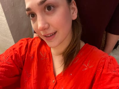 The scene is an indoor location in Ancenis, France. The primary subject is a young female, positioned in the foreground, facing the viewer with an upward and slightly rightward gaze and a subtle smile. She possesses brown eyes and hair, and is attired in a bright red, long-sleeved top featuring eyelet embroidery and a button-front V-neck. A thin gold-tone necklace is present on her neck. Partially visible behind and to the right of the primary subject is another individual, wearing a dark red garment. The background consists of a textured, light-colored wall on the left, with minimal detail visible elsewhere.