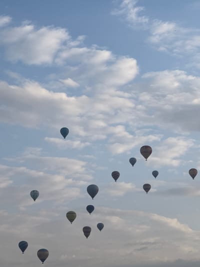 The image depicts numerous hot air balloons floating in the sky above Göreme, Turkey. The sky is light blue with scattered white clouds. The hot air balloons are various colors and patterns.