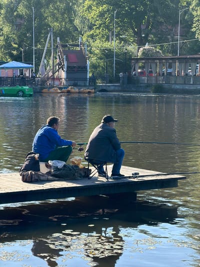 Two men are fishing from a wooden pier on a body of water. The man on the left is wearing a blue hooded sweatshirt and green pants, and is sitting with his legs crossed, holding a fishing rod. He has a bag and a white bucket next to him. The man on the right is wearing a dark jacket and a baseball cap, and is sitting on a folding chair with his fishing rod extended over the water. In the background, there is a green paddleboat on the water, a building with a blue awning, a playground structure, and a covered outdoor seating area with cars parked behind it. Trees and other vegetation line the opposite bank. The water reflects the sky and surrounding environment. The scene appears to be in Svetlogorsk, Russia.