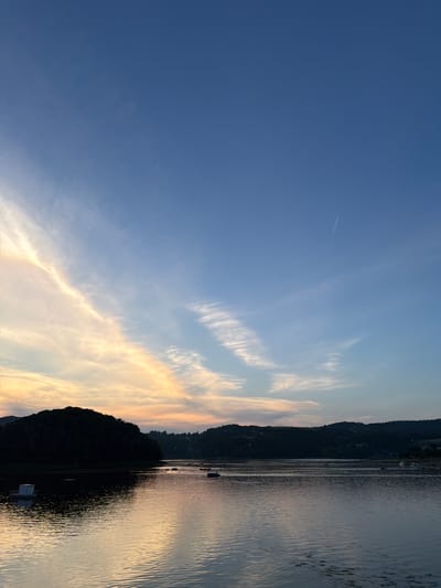 This is a landscape photograph of a body of water at sunset, likely in Gródek nad Dunajcem, Poland. The sky is a gradient of deep blue at the top, transitioning to lighter blues and pale yellows and oranges near the horizon, indicative of the sun setting. Wispy clouds stretch across the sky, catching the sunset colors. A faint airplane contrail is visible in the upper right quadrant of the sky.

The water in the foreground reflects the colors of the sky, creating shimmering patterns. Several small boats, possibly rowboats or kayaks, are scattered across the water. A white floating structure, appearing to be a small raft or buoy, is present on the left side of the frame.

In the background, a dark, silhouetted hill or mountain range dominates the left side of the image, with houses or buildings dotted along its slopes and further across the horizon. The water extends to meet the land in the distance. The overall scene is serene and captures the tranquil beauty of the natural environment at dusk.