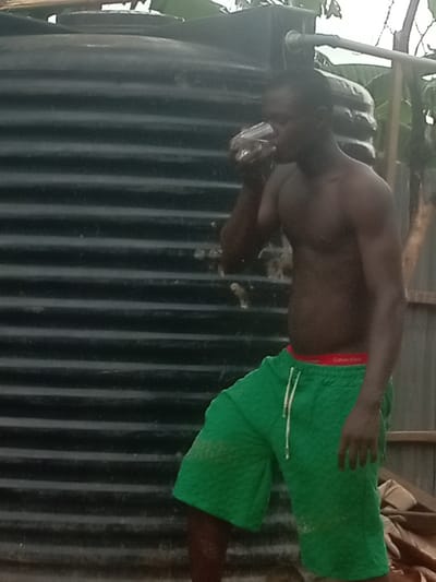 The image displays a shirtless man drinking from a clear cup outdoors, set against a backdrop featuring a large water storage tank. The man, with dark skin and a lean, muscular build, is seen from the chest down to just above his knees. He wears vibrant green shorts with a textured, possibly quilted, pattern and a white drawstring. A red elastic waistband peeks out from above his shorts, clearly displaying the brand name "Calvin Klein" in white text.
He is positioned to the right of a prominent black, corrugated water tank, which occupies a significant portion of the left side of the frame. A pipe or similar structure extends from the top of the tank into the upper right background, suggesting a water collection or distribution system. The man is focused on drinking, with a neutral expression. In the background, there is leafy green foliage, possibly banana leaves, and some indistinct structures that could be part of a fence or a building. The lighting is diffused and appears to be daytime, possibly under an overcast sky, with no harsh shadows. The scene evokes a moment of refreshment in an outdoor, semi-rural or residential setting, consistent with daily life in locations like Ihiala, Nigeria, where water storage solutions are common.