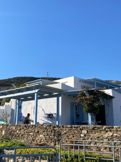 A daytime scene in Απολλωνία, Greece, depicts a white-plastered building complex with light blue wooden pergolas and shutters, set against a clear blue sky and a distant green hillside. In the foreground, a low, dry-stone wall separates the viewer from the building. Two individuals are present: on the left, an adult with dark hair, wearing a dark top, stands near an outdoor shower fixture, bending over a light-colored bucket or container. To the right, another adult, dressed in a blue shirt and dark shorts, is seated on a chair under a pergola, facing left. A dark garment is hanging on the white wall between the two individuals. A small tree with pink flowers grows beside the building on the right. A metal railing is visible in the immediate foreground, with green foliage behind it. Power lines and a utility pole are faintly visible on the distant hillside.