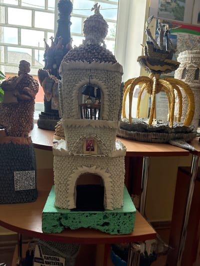 The image shows a collection of elaborate origami sculptures displayed on a wooden table near a window. The central sculpture is a white, multi-tiered tower with arched openings, a bell at the top, and a small religious icon. To the left of the tower is a dark, textured sculpture of a standing figure. Behind these, partially visible, are other origami structures including a tall, dark, spired tower and a yellow, crown-like structure with tentacle-like elements. The background features a window with a grid of panes, some vegetation visible outside, and posters on the wall. The table itself is a light wood with a reddish edge, and metal legs are visible supporting it. The overall impression is an indoor display of handcrafted art. The context of "Ветрынскі сельскі Савет, Belarus" suggests this display is likely within a cultural center, school, or community space in that Belarusian locality.