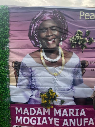 A large, creased banner, oriented vertically, dominates the frame, featuring the vibrant image of a smiling woman. The woman, depicted from the waist up, wears a purple headtie (gele) and traditional attire, adorned with a white and yellow beaded necklace and earrings. Her expression is warm and welcoming.

The banner is a commemorative piece, indicated by the visible text and decorative elements. To the top left, the word "Peace" is partially visible in a large, light-colored font. Further down, other partial words like "moth" (likely "mother"), "ERH", and "DO" (likely "daughter") can be seen. A prominent dark red strip on the right side of the banner displays the full name "MADAM MARIA MOGIAYE ANUFA" in bold white capital letters.

Attached to the banner are artificial flower arrangements: a cluster of white and green flowers near the top left, and a bunch of yellow flowers towards the bottom right. The bottom edge of the banner is lined with a strip of artificial green foliage, resembling a hedge. Above the banner, a cloudy daytime sky is visible, suggesting an outdoor setting. The creases in the banner material indicate it is made of a flexible fabric like vinyl, and has likely been rolled or folded. The overall presentation suggests a memorial service, funeral, or celebration of life for Madam Maria Mogiaye Anufa in Aghalokpe, Nigeria.