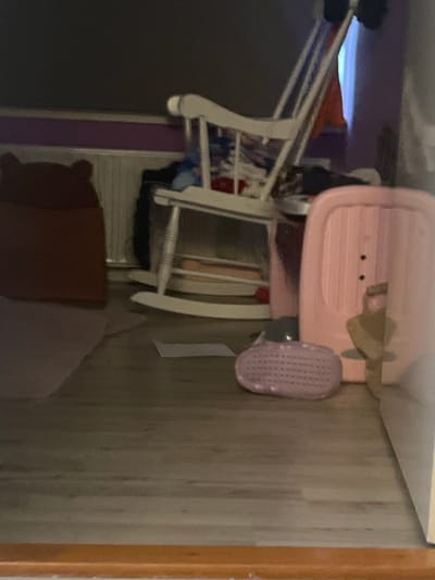 The image shows an indoor scene, likely a bedroom or living area. A pink baby tub is the most prominent object, positioned on top of what appears to be a white rocking chair. The tub has a ribbed texture and two small black dots that could be drainage holes or decorative elements. Attached to the side of the tub is a small, oval-shaped object with a grid pattern, possibly a washcloth or sponge. A tan-colored ribbon or bow is also attached to the tub. The rocking chair has white legs and a white frame with horizontal slats. Part of a brown-and-purple cushion or blanket is visible at the bottom of the frame, resting on the floor. The background consists of a wooden floor with a light-colored grain and a vertical wooden trim on the right side of the frame. A purple-colored wall or panel is visible behind the rocking chair. The lighting is dim, suggesting it might be daytime with the lights off or evening. There is no visible text in the image. The scene is static, with no activity taking place.