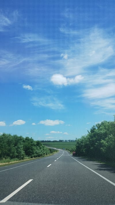 The image depicts a wide, curving asphalt road with a white dashed line and a solid white line marking the lanes. The road is bordered by lush green trees on both sides, with a glimpse of a field beyond the trees on the left. The sky is a vibrant blue with scattered white clouds, suggesting a clear day. The perspective is from a moving vehicle, giving a sense of travel along the road. The sun appears to be shining, casting shadows from the trees. The image conveys a feeling of a scenic drive through a rural or semi-rural landscape. There is no visible text in the image. The scene suggests it is daytime, likely during spring or summer, given the abundant greenery.