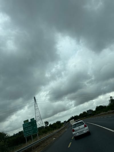 The image shows a cloudy sky over a highway in Slieverue, Ireland. A green road sign indicates directions to Rosslare, Loch Garman, Wexford, and New Ross, with distances listed in kilometers. A tall electricity pylon stands to the right of the sign. Several cars are on the highway, including a silver Volkswagen Polo with license plate 171-D-64787. The highway is bordered by trees and vegetation.