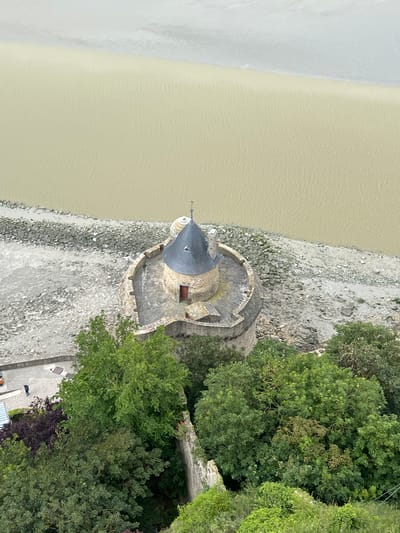 This image provides an aerial view of a circular stone structure with a conical roof, situated on a rocky shoreline adjacent to calm, muddy water. The structure appears to be a small tower or part of a larger fortification, characterized by its textured stone walls and a dark grey roof. A small wooden door is visible on the side of the tower. Surrounding the tower and extending inland is a dense cluster of vibrant green trees, suggesting a natural, possibly forested environment. The ground near the tower is covered in rocks and gravel, leading down to the water's edge.

Further inland, a paved pathway is visible, with two individuals appearing to be walking or standing on it. The presence of these people and the pathway suggests a developed area within a natural landscape. The lighting in the image is somewhat muted, indicating it might be overcast or late in the day. The overall scene evokes a sense of historical architecture set against a natural backdrop. The water appears still, with a slightly murky or sediment-laden quality. The environment seems serene and somewhat remote. No visible text is present in the image.