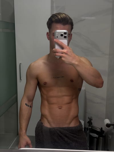 A male individual, appearing to be an adult, is captured in a mirror selfie within what appears to be a modern bathroom. His face is obscured by a smartphone, held in his right hand, which features a clear case and a prominent triple-lens camera array. The individual has light brown hair with subtle highlights, styled back. He exhibits a muscular physique, revealing his torso with a visible six-pack. A dark grey towel is wrapped around his waist, covering his lower body.
Distinguishing features include:
- A tattoo on his upper chest, centered, displaying the word "IMPERFECT" in reverse due to the mirror reflection.
- Two barbell nipple piercings.
- A tattoo of vertical script, consistent with East Asian characters, on his left forearm.
- A partially visible tattoo on his right torso, near the hip.
- A small earring is visible in his left ear.
The bathroom environment, reflected in the mirror, features light-colored, possibly marble-patterned, tiled walls. Visible fixtures include a white toilet with its cistern, a toilet paper holder with a roll of paper, a metallic toilet brush holder, and a bidet spray hose mounted on the wall. A glass shower partition or cabinet with a metallic handle is visible on the left side of the frame. The lighting appears even, suggesting indoor artificial illumination.
The image provides no discernible features or landmarks that permit identification of its specific location within the city of Viseu, Portugal, or any other geographical area.