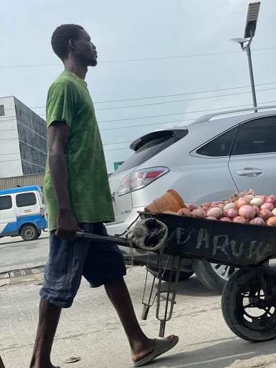 In Ojomu, Nigeria, a Black man with dark skin and short, curly hair is captured in a profile view from the waist up, walking to the right. He is wearing a green t-shirt with a tie-dye pattern and dark blue shorts. He is pushing a black metal cart filled with onions and a small orange bucket. The cart has wheels and the word "OHURU" is faintly visible in white lettering on its side. Behind the man and his cart, a silver SUV is parked, and further back, a white and blue van is visible. The scene takes place outdoors on a paved surface, with power lines overhead and a modern building in the background. The sky is overcast.