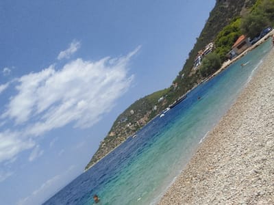 The image shows a coastal scene in the Municipal Unit of Solygeia, Greece. A steep, tree-covered hillside slopes down to a pebble beach and the clear blue-green waters of the sea. Several houses are visible nestled into the hillside. The sea is calm, with a few boats anchored offshore. A person is swimming in the water. The sky is blue with scattered white clouds. The perspective is from a low angle, looking up at the hillside.