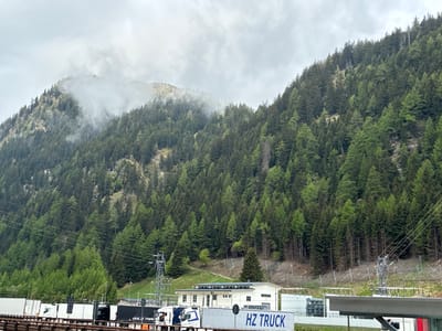 The image shows a view in Brenner - Brennero, Italy. A forested mountain slopes upwards towards the left and right, partially obscured by low-hanging clouds at its peak. The mountain's slope is densely covered with green coniferous trees, with slight variations in color suggesting different tree types or ages.
In the foreground, a transportation area is visible. A series of trucks are lined up, some bearing the "HZ TRUCK" logo. A white building with a dark roof is situated behind the trucks, near power lines and electrical equipment. The scene suggests a transportation hub located in a mountainous, forested area. The weather appears overcast, as indicated by the cloud cover and the overall muted lighting.