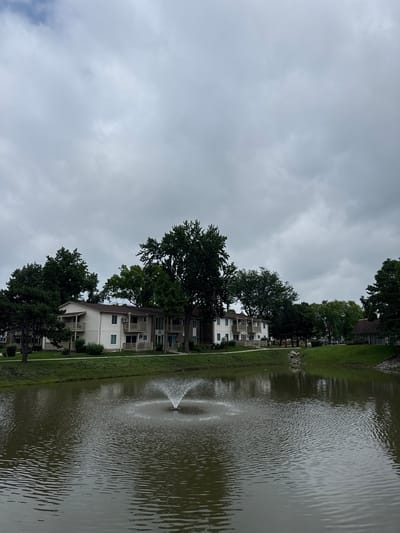 The image depicts a tranquil scene in Woodridge, United States of America, featuring an apartment complex situated beside a pond. The sky above is overcast with thick, gray clouds, casting a subdued light on the environment. In the foreground, the pond's surface is disturbed by ripples, with a fountain actively spraying water in the center, creating a delicate mist.
The apartment buildings are low-rise structures, painted in a light beige color, with multiple balconies and windows visible. They are nestled amongst a variety of trees, some of which are tall and leafy, providing a natural backdrop. The grounds are well-maintained, with green grass covering the slopes leading down to the pond. The overall impression is one of a peaceful residential area, perhaps an apartment community, with a focus on natural elements and communal spaces. The scene appears to be devoid of any human presence at the time the photograph was taken.