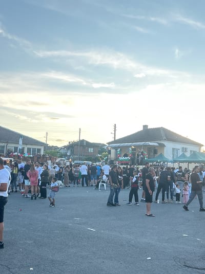The image depicts an outdoor gathering in Gabar, Bulgaria, likely a community event or festival. The scene is populated by a large crowd of people, including adults and children, engaged in various activities. In the foreground, several individuals are standing and walking, some appearing to interact with each other. A young boy in the middle ground is holding what appears to be a toy guitar. In the background, buildings, tents, and decorations such as balloons can be observed. The setting suggests a festive atmosphere, possibly a street fair or celebration, taking place in what seems to be a residential or village area. The sky above is partly cloudy, indicating it is daytime.