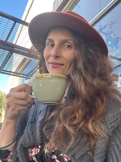 A woman, wearing a rust-colored hat, a gray knitted cardigan over a black floral dress, and a silver bracelet on her left wrist, holds a light green mug with a gold rim in her right hand to her lips. She is outdoors, with a clear blue sky overhead and a lattice-like structure behind her, suggesting an outdoor seating area. The background includes a large window reflecting the sky and some industrial elements. To the left, palm fronds are visible. The woman has long, wavy brown hair and is looking directly at the camera with a slight smile.