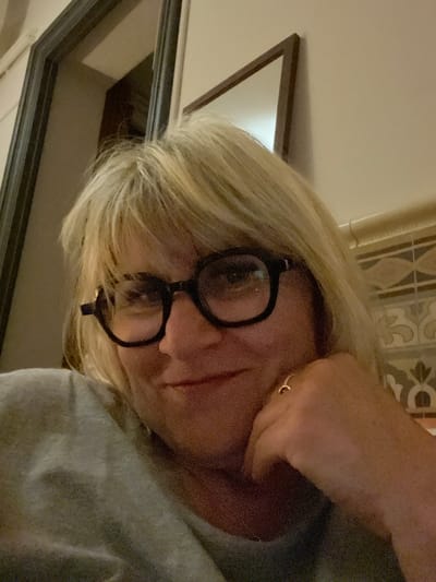 A medium close-up angled shot depicts a woman with light blonde hair wearing round, black-rimmed glasses. Her right hand is raised to her face, touching her cheek. She has a small gold ring on her finger and a small gold earring. She is wearing a gray sweater. In the background, there is a framed mirror and decorative tile work. The location, Montélimar, France, cannot be determined from the image alone.