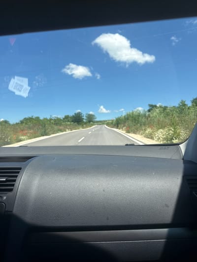 The image captures a view from the interior of a vehicle, looking through the windshield at a road stretching into the distance. The road is paved and marked with white lines, suggesting it is a country road. On either side of the road, there is lush green vegetation, with some patches of red flowers and white wildflowers. The sky is a clear blue with a few fluffy white clouds.

The interior of the car is visible in the foreground, with a dark gray dashboard. On the windshield, there are two small white stickers with QR codes. The overall impression is a sunny day, likely in a rural setting outside of the city of Suvorovo, Bulgaria, given the vegetation and the open road. There are no people visible in the image.