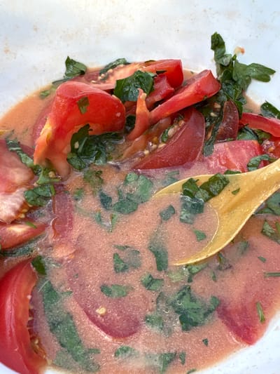 The image shows a close-up of a bowl of gazpacho, a cold Spanish soup made from raw, blended vegetables. The soup is predominantly red, with chunks of fresh tomatoes and finely chopped green herbs, likely basil or parsley, visible throughout. A wooden spoon is partially submerged in the soup, suggesting it's being prepared or served. The soup has a slightly textured, pulpy appearance with small bubbles on the surface, indicating it might be chilled or recently blended. The overall setting appears to be a kitchen or dining area, with the bowl likely placed on a table or countertop. The lighting is natural, suggesting daytime, and there are no obvious external cues for weather. The focus is entirely on the food, emphasizing its freshness and vibrant colors. No text is visible in the image.