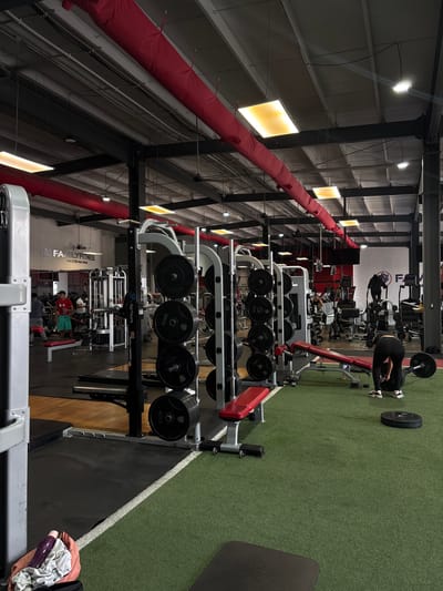 The image captures the interior of a Rayon Family Fitness gym, located in San Pedro Cholula, Mexico. It presents a view of the gym equipment and interior architecture. The layout includes weight racks, exercise machines, and free weight equipment such as barbells and plates. A person is visible using equipment near the weights. The gym features an industrial aesthetic, with exposed steel beams supporting the structure. Overhead, there is a prominent red ductwork system along the ceiling. The floor is covered with artificial turf and lined with white and black sections. The logo of "Rayon FAMILY FITNESS" is visible on the wall.