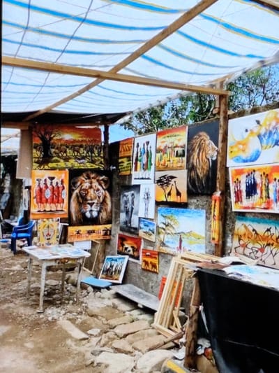 This image depicts an outdoor art market stall in Ihumwa, Tanzania. The scene is illuminated by natural light, showing a collection of paintings displayed under a makeshift canopy made of wooden beams supporting a white and blue striped fabric roof. The ground surface consists of packed dirt and scattered light-colored stones.
Numerous colorful paintings, primarily on canvas, are hung directly on a rough, grey wall that forms the back of the stall, with additional pieces leaning against the wall or stacked on the ground. The artworks predominantly feature African themes, including detailed portraits of wildlife such as lions (one prominent frontal portrait, one in profile), elephants, giraffes, and rhinos. Other paintings depict scenes of Maasai people in traditional attire, vast savanna landscapes with acacia trees and sunsets, and some abstract interpretations of African motifs.
In the mid-ground, a rectangular white wooden table stands on uneven legs, holding a display of smaller artworks, postcards, or prints. To the left of this table is a single blue plastic chair. On the right side of the frame, a stack of unpainted wooden stretcher frames or canvases leans against the wall, alongside other items covered by dark fabric. Green foliage and a bright sky are visible above the structure in the background. No individuals are present in the image.