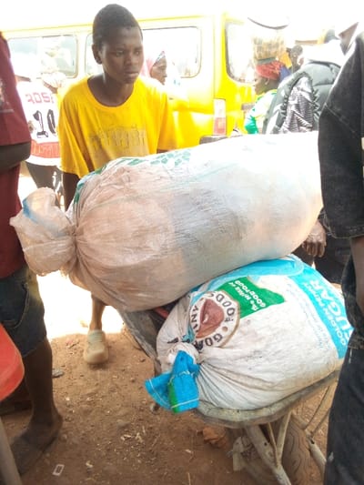 The scene is located in Gana Daji, Nigeria, depicting an outdoor market or street environment. A young male, wearing a yellow t-shirt and light-colored clogs, stands centrally, positioned behind two large, stacked sacks. The upper sack is an elongated, off-white woven bag containing bulk material, tied at one end. This sack rests on a lower, larger white woven bag. The lower bag displays "GOAT FOODS" within a brown circular logo and "Golden Penny SUGAR" in blue and white text; it is tied with a blue and green cloth and rests on the bed of a metal wheelbarrow, of which a wheel is partially visible.
To the left, a partial figure wears a dark red t-shirt with a white graphic and dark shorts. To the right, another partial figure wears dark clothing. In the background, several individuals are present; a woman with a light head covering is visible to the left of the central figure, and another woman with a red head covering and green top is visible further right. A bright yellow van or bus is parked in the background, and a red container sits near the vehicle. The ground is unpaved reddish-brown dirt. The scene is brightly lit by daylight.