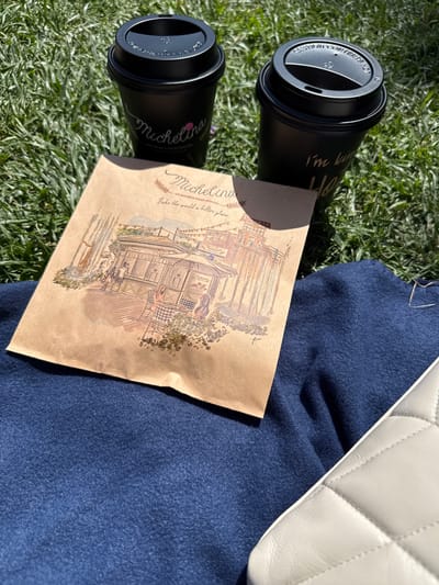 Two black coffee cups with lids rest on a patch of green grass. One cup has the word "Michelina" in cursive and "ARTISAN BOULANGER-PATISSIER" written below it. The other cup has "I'm here" written in cursive. Between the cups is a brown paper bag with an illustration of a quaint building with people sitting at tables and a statue. The paper bag has the word "Michelina" in cursive and "Bake the world a better place" below it. To the right of the paper bag, a dark blue fabric is visible, and partially in the bottom right corner is a white quilted object that appears to be a handbag. The scene is lit by sunlight, casting shadows on the grass. The image appears to have been taken outdoors in Glendale, United States of America.