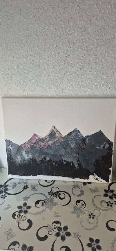 The image shows a canvas painting of mountains, with a dark, textured wall in the background and a patterned surface beneath the canvas. The painting is abstract, with thick brushstrokes of gray, black, and hints of pink and blue representing the mountain peaks and a forest at the base. The patterned surface appears to be a tablecloth or a decorative covering, featuring swirling black designs and floral and butterfly motifs with a reflective quality. The wall has a rough, stippled texture. The overall impression is that the painting is displayed on a table or surface in a domestic setting. There is no indication of the city of Baden-Baden, Germany, in the image.