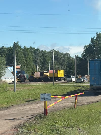 The image depicts an outdoor scene in the city of Zarech'e, Russia, on a sunny day. In the foreground, a dirt road is visible, partially obstructed by a red and yellow striped barrier. To the right of the barrier, a sign is posted, which appears to be informational or cautionary, though the text is illegible in the provided image.

The midground reveals a grassy area leading to a collection of vehicles and structures. A red tractor is parked to the left, and to its right, a large yellow trailer loaded with wooden beams is situated. A white van is also present near the tractor. Further to the right, there are other vehicles, including a dark-colored car. A blue shipping container is positioned on the far right of the image.

The background is dominated by a dense line of green trees under a clear blue sky with scattered clouds. Power lines are strung across the scene, supported by utility poles. The overall impression is that of an industrial or agricultural area.