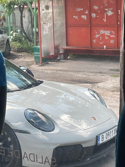 A white Porsche car is the central subject of this image, parked outdoors during what appears to be a bright, sunny day. The car, viewed from the front-right side, displays a visible Indonesian license plate, "B 0911 XM," on its front. The license plate format indicates registration in Jakarta, Indonesia.

On the lower side of the car's front fender, bold, inverted text is visible, reading "SALIDA DE." This Spanish phrase translates to "Exit of" or "Departure from" and appears to be a custom decal or part of a livery on the vehicle.

In the background, a large reddish-orange metal gate or fence is prominent, adorned with remnants of torn posters or papers. To the left of the gate, there is some foliage and a green container or bin with partially visible, unreadable text. The ground surface appears to be asphalt or concrete. The image is captured from inside another vehicle, with parts of its dashboard and windshield visible at the bottom and top of the frame, respectively. No individuals are present in the scene.