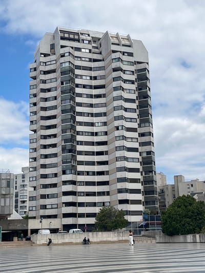 The image shows a tall, modern apartment building with a distinctive stepped facade in Créteil, France. The building is constructed with white and beige panels, and features numerous windows and balconies. The sky is partially cloudy. In the foreground, a paved public space with a striped pattern is visible. Several people are present, including a woman in white pants walking away from the camera, a man sitting on a low wall, and two individuals in a wheelchair. The background includes other buildings and trees. The scene appears to be a public square or pedestrian area adjacent to the apartment building.