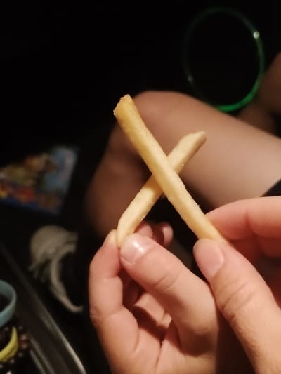 In the foreground of the image, a person's hands are holding two French fries, forming an "X" shape. The French fries are golden brown and appear to be salted. The background is dark, with a blurred green circle on the right side, possibly a glow stick or bracelet. To the left, there's a hint of a blurred object, potentially a bag or container. The scene appears to be indoors, possibly in a dimly lit setting like a car or a room at night. There is no identifiable location in the city of Gorki-2, Russia.