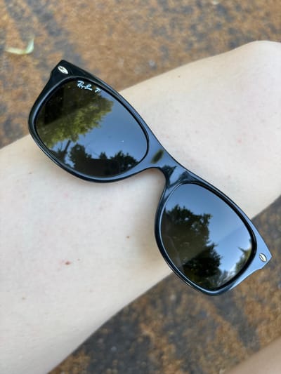 A pair of black Ray-Ban sunglasses with polarized lenses lies on a person's leg. The sunglasses are resting on skin that has some small red marks and freckles. The lenses reflect trees and the sky. The background is blurred and appears to be a textured outdoor surface. There is no indication of the location within Erlenbach am Main, Germany.
