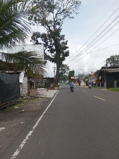 The image depicts a daytime scene on a paved road in Khajuripada, India. The asphalt road features a solid white line on its left edge and a broken yellow line indicating the center. A single individual on a motorcycle, wearing a blue shirt, is visible traveling away from the camera in the left lane. A second motorcycle is parked or slowly moving on the right side of the road further in the distance.
On the left side, the road transitions to a dirt shoulder with scattered debris, including a yellow spherical object. This leads to a narrow concrete sidewalk. Adjacent to the sidewalk are several structures, including a partially obscured white signboard, dense palm fronds, and a building section covered by a black tarp. A small, round brick structure and an orange traffic cone are also present on the left. A prominent, tall deciduous tree stands beside the road, its branches extending upwards. Numerous utility poles and overhead power lines crisscross the sky.
The right side of the road also features commercial or residential structures, with some signage visible in the distance. The background reveals additional trees and distant hills under an overcast sky.