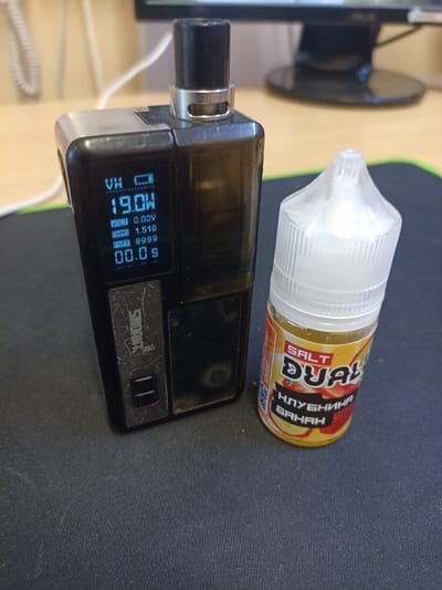 The image shows a close-up of electronic smoking devices and e-liquid on a dark, textured surface, likely a desk mat. On the left is a black electronic cigarette device with a metallic top. Its digital screen displays "VW," a battery indicator, "19.0W" for wattage, "VOLT 0.00V," "OHMS 1.51Ω," "PUFF 9999," and "00.0 s." The brand name "SMOANT" is visible on the device. To the right of the device is a clear plastic bottle of e-liquid with a white cap. The label on the bottle is primarily yellow and orange, featuring the words "SALT" and "DUAL" prominently. Below these, in Cyrillic script, it reads "КЛУБНИНА БАНАН," which translates to "Strawberry Banana." Some text in a smaller font is also visible on the left side of the e-liquid label, possibly indicating ingredients or warnings. In the background, a computer monitor and a coiled white cable can be partially seen, suggesting an indoor setting, possibly a home office or workspace. The lighting appears to be artificial, and there are no indications of time of day or weather conditions. The focus is clearly on the vaping products.