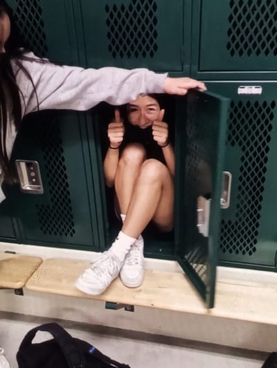 The image displays an indoor locker room or changing area, although specific identifying features for Aktash, Uzbekistan, are not present. A young female individual is seated inside an open, dark green metal locker, positioned centrally. She is smiling at the viewer, wearing a dark top and shorts, and giving a double thumbs-up gesture. Her legs are crossed at the ankles and rest on a light-colored wooden bench that spans the locker units below. She wears white crew socks, one displaying a red Nike swoosh, and white Nike athletic shoes. Multiple dark wristbands are visible on her wrists. Partially visible on the left is another young female, wearing a light gray hooded long-sleeved top. Her left arm extends horizontally, with her hand resting on the locker structure above the first subject's head. A dark-colored backpack is situated on the floor in the lower-left foreground. The lockers feature perforated ventilation patterns.