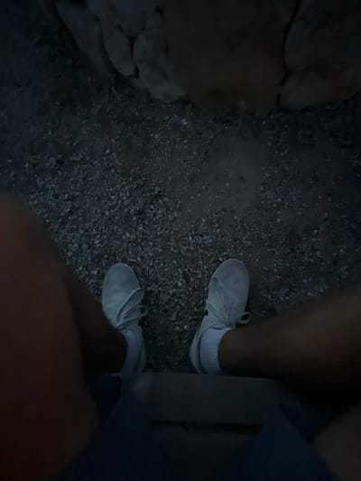 The image shows a person's legs and feet, wearing white sneakers with black accents and white socks, seated on a dark, rough surface. The person is facing the camera, with only their legs and feet visible. The background is a dark, rocky surface, possibly a rock wall or a pile of rocks. The image appears to have been taken at dusk or in low light conditions, as the environment is poorly lit. The image was taken in Paso Robles, California, specifically at the coordinates 35.608° N, 120.725° W.