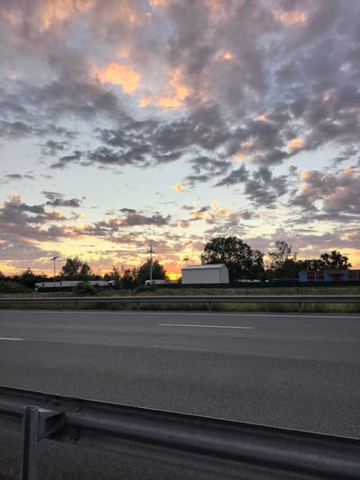 A twilight sky with dramatic clouds, painted in hues of orange, pink, and gray, forms the backdrop for this scene. The sun is setting just above the horizon, casting a warm glow through the clouds. In the mid-ground, a row of white semi-trailer trucks are parked along a road, some with visible logos. Several trees are scattered behind the trucks, and a tall, thin wind turbine stands to the right, its blades silhouetted against the sky. A white shed-like building is also visible on the left side of the scene, and further to the right, a building with blue siding can be seen. In the foreground, a dark asphalt road stretches across the frame, with a metal guardrail running along its edge. The environment appears to be a roadside rest stop or industrial area on the outskirts of Saint-Aybert, France.