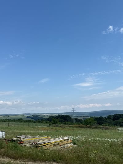 The image depicts a bright, sunny daytime landscape in Bliznatsi, Bulgaria. The upper two-thirds of the frame are filled with a clear blue sky, adorned with scattered, wispy white clouds.
The lower right portion of the image shows a vibrant green field, teeming with wildflowers, primarily small white blossoms and occasional red poppies. Resting on the edge of this field is a stack of construction materials, appearing to be wooden and metal formwork or scaffolding. Visible text on some of these stacked pieces includes "STRADYVENT".
Further into the landscape, a large metal electricity transmission pylon stands in the mid-distance, near a distant line of dark green trees, which likely marks a forest or hillside. Beyond the trees, the terrain extends into lighter green fields and rolling hills, fading into a hazy horizon. Power lines are faintly visible stretching across the sky.
The scene suggests a peaceful, rural or semi-rural environment on a clear day. No specific human or animal activity is overtly taking place within the frame. The overall impression is one of natural expanse under bright daylight conditions.