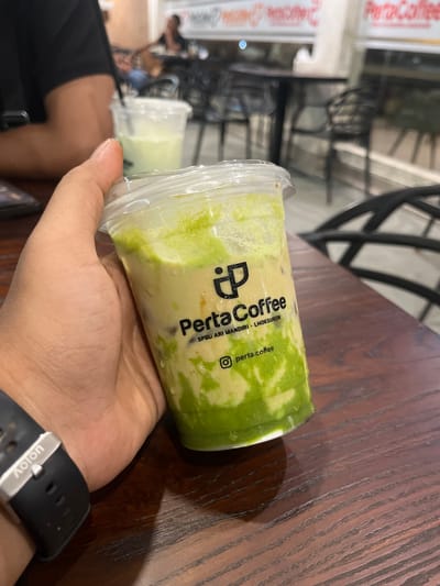 A close-up, eye-level shot shows a person's hand holding a clear plastic cup of a green, layered beverage. The cup has a black lid and a straw. The beverage appears to be a matcha drink with visible layers of green syrup and creamy liquid, with ice. A black smartwatch with a silver buckle and the word "Aolon" is visible on the person's wrist. The cup has a logo and text printed on it. The logo is a stylized 'P' inside a square. Below the logo, text reads "PertaCoffee" in black, and below that, "SPBU ARI MANDIRI - LHOKSUKON". At the bottom of the cup, text reads "p pta.coffee" with an Instagram logo. The background is out of focus, showing a cafe interior with tables, chairs, and other patrons. The setting is likely a coffee shop in Meunasahceubrek, Indonesia, given the text on the cup.