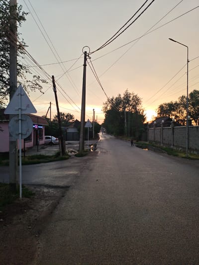 This image captures a street scene in Іргелі ауылдық округі, Kazakhstan, during what appears to be twilight. The sky is a pale orange and yellow gradient, indicating either sunrise or sunset. Overhead, numerous electrical wires crisscross the frame, supported by utility poles. A street lamp is visible on the right side of the road.
The road itself is paved and wet, with several puddles reflecting the dim light. The left side of the road features a small, pink building with a dark awning and windows, possibly a shop, with a silver car parked in front. A white triangular road sign with a red border is prominent in the foreground on the left. Further down the street, there are more utility poles and a long, grey stone fence bordering the right side of the road. Trees with dark foliage line the street, silhouetted against the colorful sky. A lone figure, appearing to be a person, is walking away from the camera on the right side of the road. The overall impression is a quiet, somewhat rural street at the end of the day.