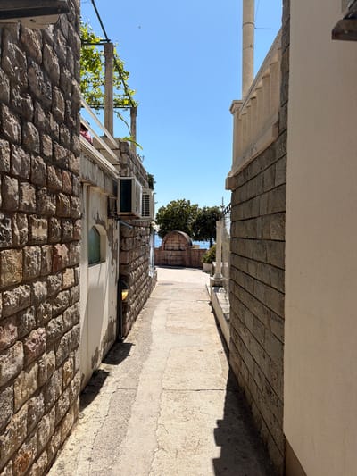 The image captures a narrow, sunlit alleyway between two buildings in what appears to be a coastal town, consistent with the provided location of Nerin, Montenegro. The perspective looks down the passage towards a distant view of the sea.
On the left, a building constructed with rough-hewn stone blocks rises from the ground. Above the stone section, the wall is lighter, possibly stuccoed, and features a small arched window. An air conditioning unit is mounted on this upper wall, and a wooden trellis adorned with green vines extends towards the clear blue sky.
The right side of the alley is also lined by a building with a stone base. A light-colored concrete or paved path runs alongside this structure. Strong sunlight casts sharp, elongated shadows from the building and an unseen overhead structure across the path, indicating a bright, sunny day, likely around midday or early afternoon. The ceiling above this path is a flat, light-colored surface, suggesting a covered area or an architectural overhang.
Further down the alley, beyond the immediate buildings, a glimpse of the calm, blue sea is visible under an equally clear blue sky. Some green trees and a section of a white, decorative balustrade or pillar can also be seen, contributing to the Mediterranean aesthetic of the scene.
No people or activities are visible in the image, presenting a quiet and still urban landscape. There is no visible text. The scene is dominated by the textures of stone masonry, the interplay of light and shadow, and the vibrant blue of the sky and sea, characteristic of a picturesque coastal setting.