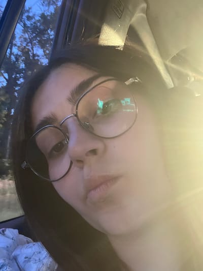 The image is a close-up selfie of a young woman wearing large, round glasses. She has dark hair and appears to be in a car, as suggested by the window frame and the interior of the vehicle visible in the background. The lighting is bright and warm, with a lens flare dominating the right side of the image. Outside the window, trees and a twilight sky are visible. The woman is looking towards the camera with a neutral expression. There is no discernible information in the image that would indicate a specific location within Luzhany, Ukraine.