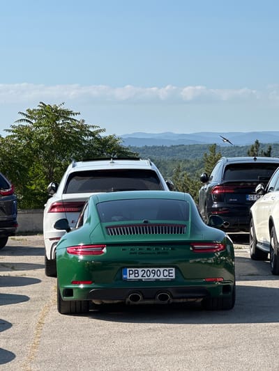The image captures a close-up rear view of a dark green Porsche 911 Carrera GTS parked in Arbanassi, Bulgaria. The car's license plate reads "PB 2090 CE" with the country code "BG" indicating Bulgaria. To the left of the Porsche, the rear of a white Audi Q8 is visible, and further left, part of a dark-colored SUV. To the right of the Porsche, the rear of a white car is partially visible. In the background, a lush green tree dominates the frame, with a mountainous landscape and a clear blue sky beyond. A bird is in flight in the upper right quadrant of the image. The setting appears to be an outdoor parking area, likely on an elevated position given the view of the landscape.