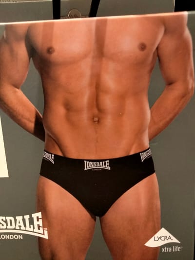 An image depicts a torso of a man wearing black briefs with a white "Lonsdale London" logo on the front and the sides. The man has visible abdominal muscles and tanned skin. A sliver of a partial "Lonsdale London" logo is visible on the left corner of the image. The lower right corner displays "LYCRA. xtra life" in a triangular shape. There is a line crossing the image from the top middle.
