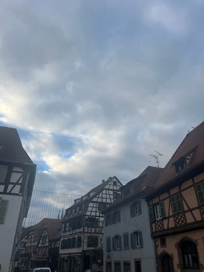 The image displays a street scene in Obernai, France, featuring a row of historic buildings under a predominantly overcast sky with visible patches of blue. The architecture is characterized by multi-story structures, many exhibiting traditional half-timbered facades with dark timber frames and lighter infill. Pitched roofs with reddish-brown tiles are prominent. Several windows with closed and open shutters are visible on the building facades. On the roof of a building on the right, an outdoor television antenna is present. Strung across the street, between the buildings, are decorative string lights composed of numerous small bulbs. No individuals or specific actions are discernible in the frame.